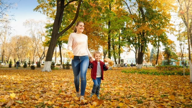 Happy laughing little boy walking his mother on yellow fallen leaves at autumn park.