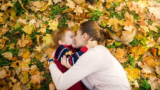 Portrait of little boy kissing his young mother lying on ground at autumn park.