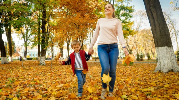 Happy smiling young mother holding her son by hand and walking at autumn park.