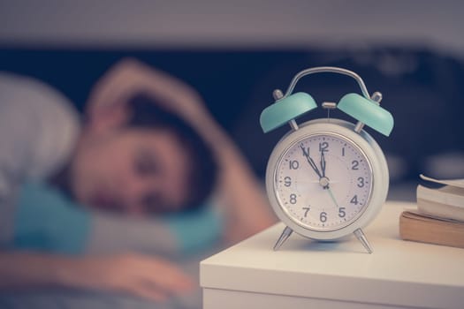 White alarm clock on the bedside table. Young man sleeps in the background.