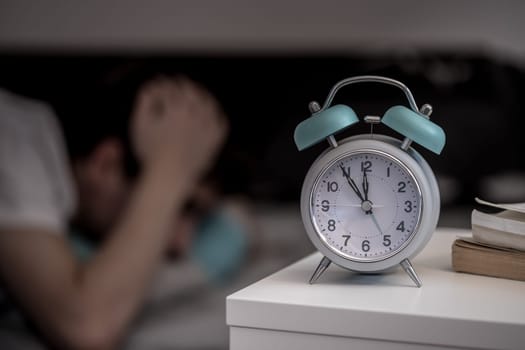 White alarm clock on the bedside table. Young tries to sleep in the background, insomnia