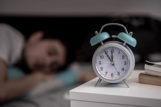 White alarm clock on the bedside table. Young man sleeps in the background.