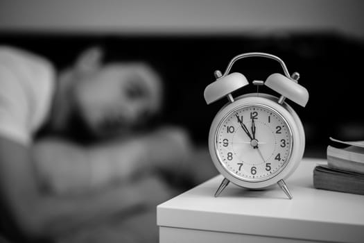 White alarm clock on the bedside table. Young man sleeps in the background.