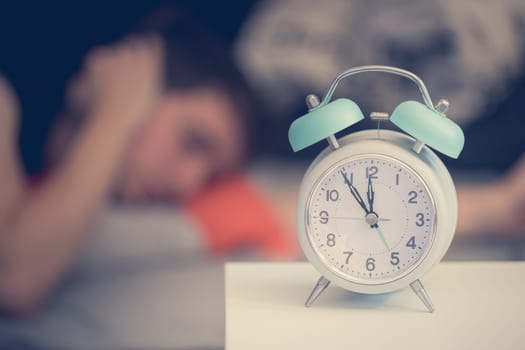 White alarm clock on the bedside table. Young tries to sleep in the background, insomnia