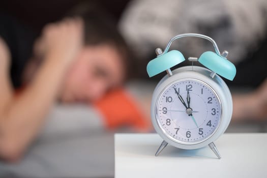 White alarm clock on the bedside table. Young tries to sleep in the background, insomnia