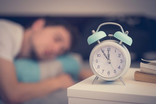 White alarm clock on the bedside table. Young man sleeps in the background.