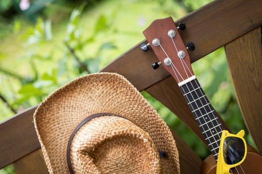 Ukulele and yellow sunglasses on a wooden park bench in summer, green area in the blurry background