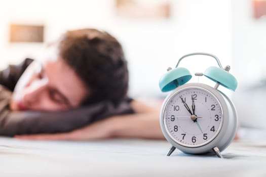 White alarm clock in the morning. Young man sleeps in the background.