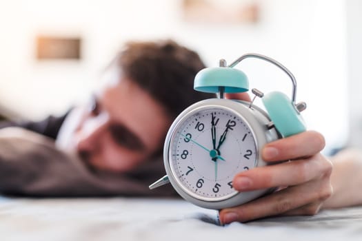 White alarm clock in the morning. Young man sleeps in the background.