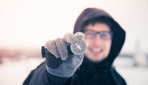 Young smiling man holds up a compass. Wintertime, gloves and hood.