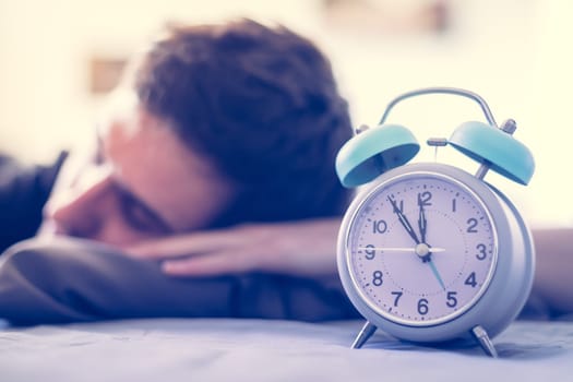 White alarm clock in the morning. Young man sleeps in the background.