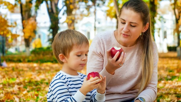 Portrait of little todler boy with mother eating red apples in autumn park.