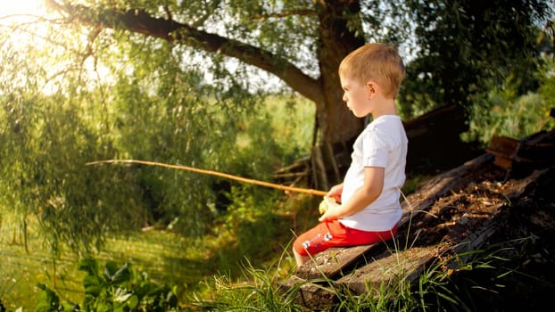 Cute little boy sitting on the bench at river and fishing with old wooden rod.