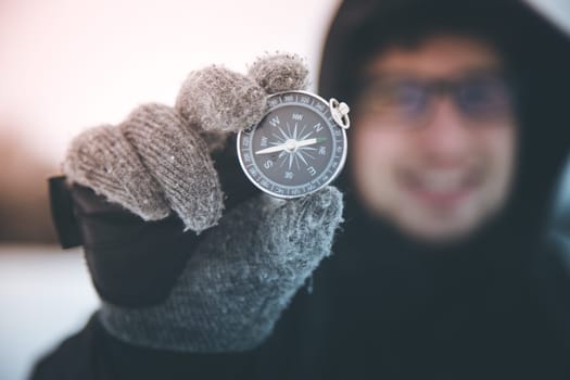 Young smiling man holds up a compass. Wintertime, gloves and hood.