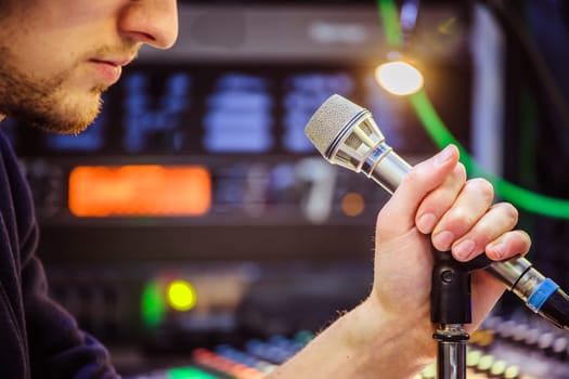 Young man is sitting in the recording studio and talks into the microphone, buttons and equipment in the blurry background