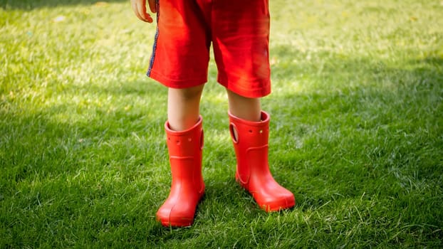 Closeup photo of child wearing red rubber boots standing on grass at park.