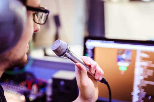 Young man is sitting in the recording studio and talks into the microphone, buttons and equipment in the blurry background