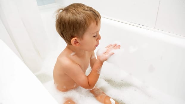 Little toddler boy sitting in bath and playing with soap foam.