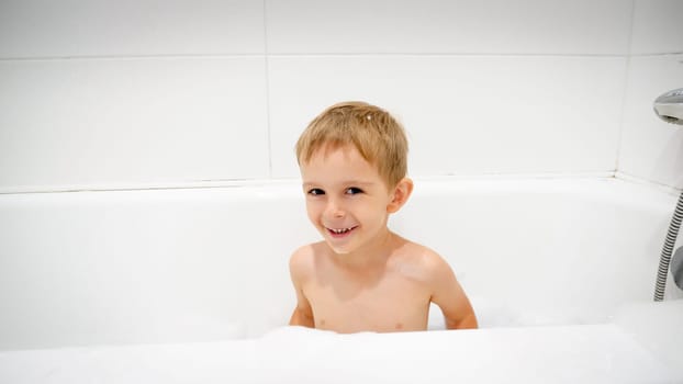 Portrait of cute smiling little boy sitting in bath full of soap foam.