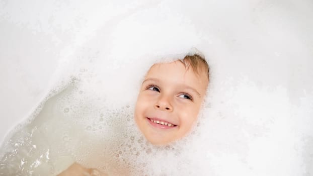 Portrait of cute smiling little boy lying in bath covered with soap foam.