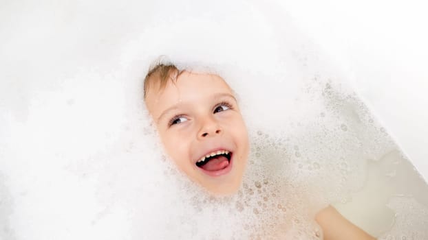 Funny portrait of cute little boy lying in bath with soap foam.