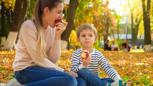 Portrait of little smiling boy with mother sitting on ground at autumn park and eating red apples.