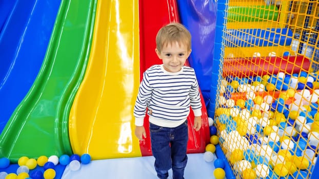 Portrait of happy smiling little boy standing in playroom with colorful slide at amusement park.
