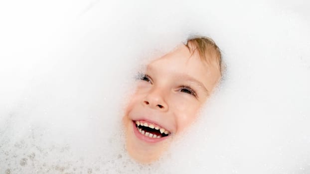 Portrait of happy laughing boy covered in soap foam swimming and playing in bath.