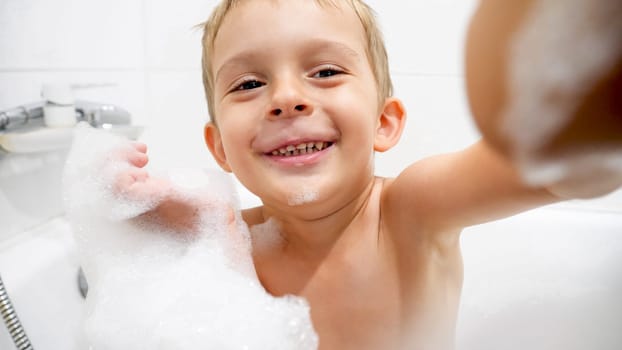 Portrait of cute toddler boy covered in soap foam taking bath and looking in camera.