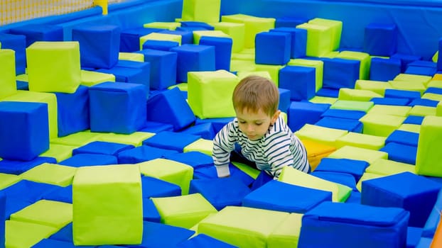 Little boy crawling and playing with soft cubes in big pit on playground at shopping mall.