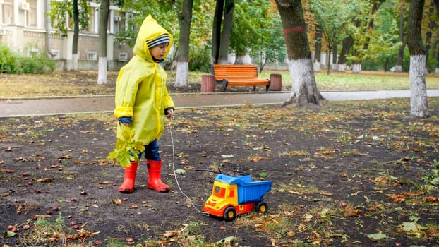 Little toddler boy in raincoat playing with toy truck at park on cold rainy day.