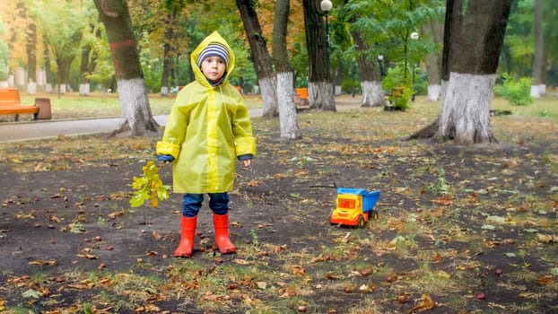 Little toddler boy in raincoat and red rubber boots at park on rainy day.