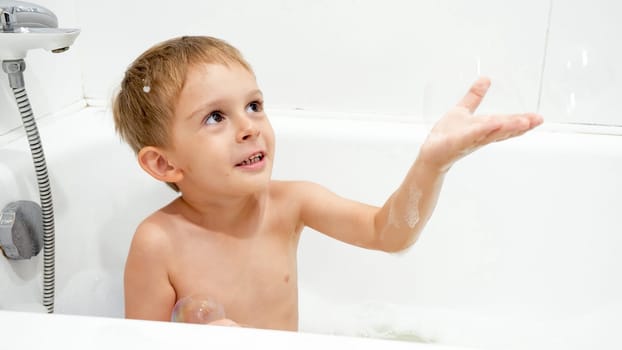 Portrait of cute boy catching soap bubble while washing in bath.