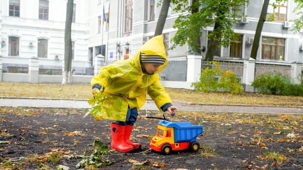 Little boy in raincoat and rubber boots ppicking up fallen autumn leaves and putting it his toy dump truck.