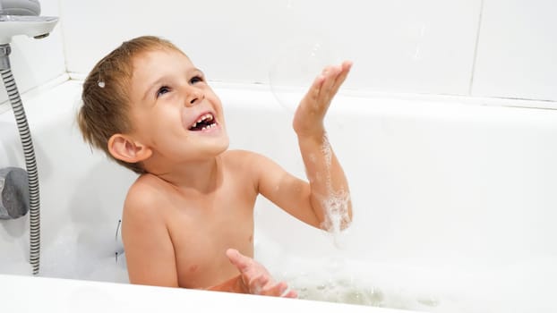 Happy smiling boy playing with soap bubbles and foam in bath.