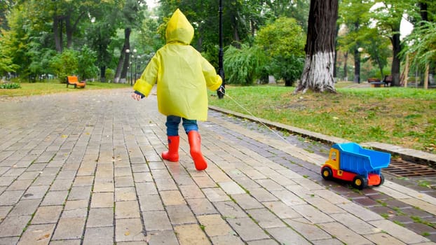 Cute little boy in yellow raincoat running with toy truck under rain at park.