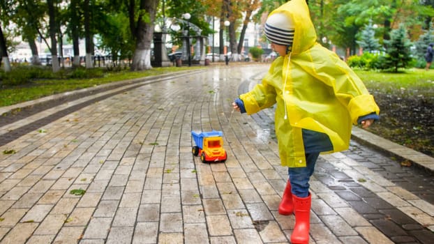 Cute toddler boy in yellow raincoat running with toy truck in rainy autumn park.