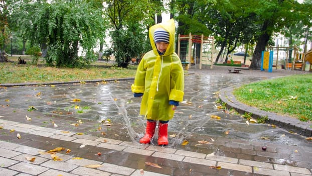 Cute little boy in raincoat and red wellington boots jumping in big puddle at park.