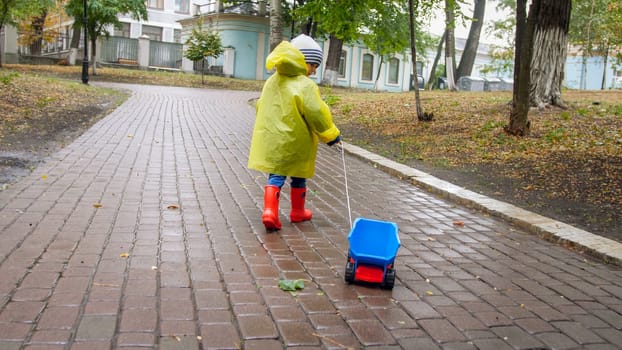 Little 4 years old boy in boots and raincoat walking at park with his toy truck.