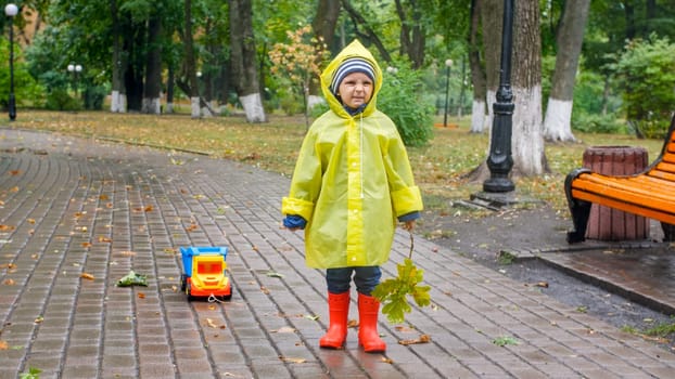 Little toddler boy in raincoat and rubber boots got upset because of bad weather and heavy rain in park.