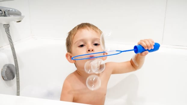 Portrait of cute little boy blowing soap bubbles while washing in bath.