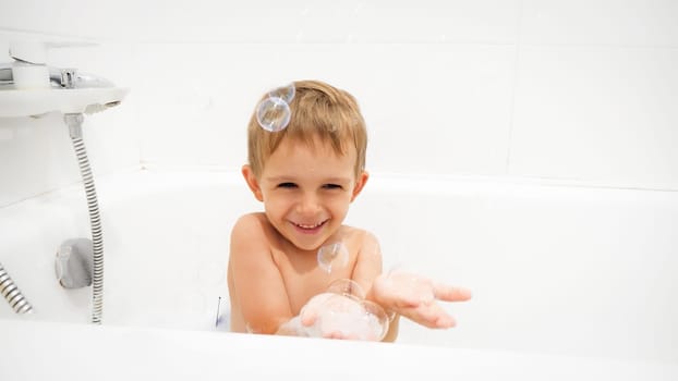 Portrait of cheerful laughing little boy washing in bath and cathing soap bubbles on hand.