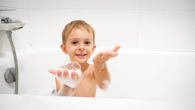 Portrait of cute toddler boy having bath cathing flying soap bubbles.