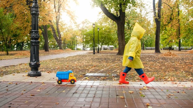 Cut little boy in red rubber boots and yellow raincoat walking with toy truck at autumn park.
