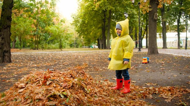Portrait of happy laughing toddler boy in yellow raincoat and rubber boots enjoying walking in autumn park on rainy day.