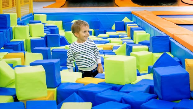Portrait of happy smiling toddler boy enjoying playing with soft cubes in big pitt in trampoline park.