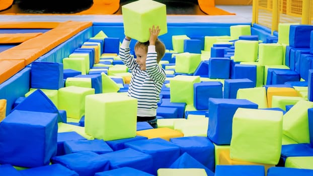 Happy smiling toddler boy playing and throwing soft cubes in big pit at playroom in mall.