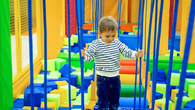Little concentrated boy balancing on ropes and bridges in children playroom at shopping mall.