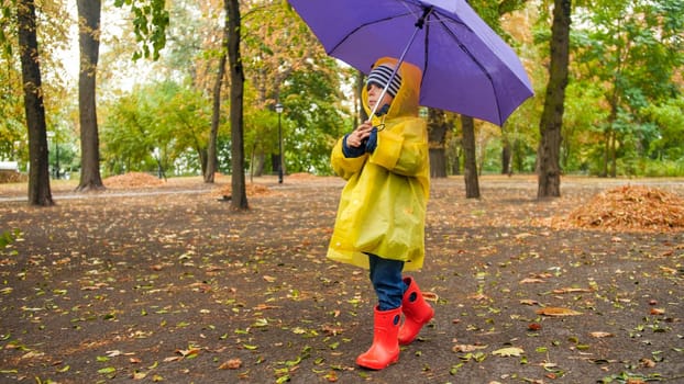 Little toddler boy in wellington booats and raincoat walking at park with umbrella.