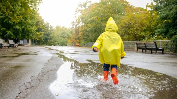 Little happy boy in rubber boots running over big puddle after rain at park.